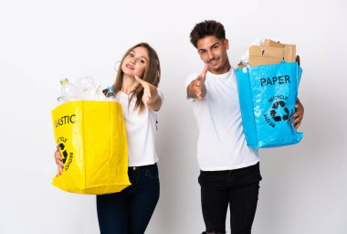 Beyaz arka planda izole edilmiş plastik ve kağıtlarla dolu bir çanta tutan genç çift iyi bir anlaşma için el sıkışıyor.