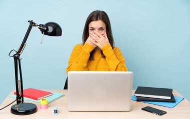 Öğrenci kız evinde çalışıyor. Mavi arka planda izole edilmiş. Ağzını elleriyle kapatıyor.