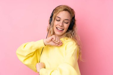 Pembe arka planda izole edilmiş sweatshirt giyen genç sarışın kadın müzik dinliyor ve dans ediyor.