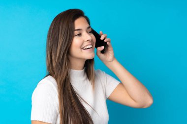 İzole edilmiş mavi arka planda genç bir kız cep telefonuyla sohbet ediyor.