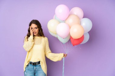 Mor arka planda bir sürü balon yakalayan genç bir kadın gözleri kapatıyor ve parmaklarından bakıyor.