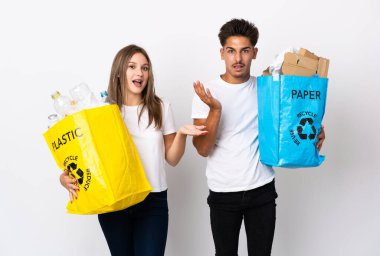 Plastik ve kağıt dolu bir çanta taşıyan genç bir çift beyaz arka planda şok olmuş bir yüz ifadesiyle izole edilmiş.