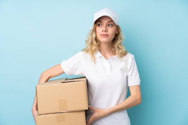 Genç teslimatçı kadın mavi arka planda izole edilmiş duruyor ve yan tarafa bakıyor.