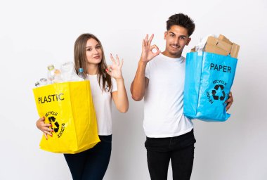 Beyaz arka planda izole edilmiş plastik ve kağıt dolu bir çanta taşıyan genç çift, parmaklarıyla işaret veriyor.