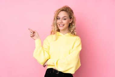 Pembe arka planda izole edilmiş eşofman üstü giyen genç sarışın kadın parmağıyla kenarı işaret ediyor.