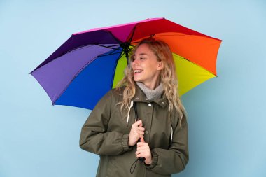 Mavi arka planda şemsiye tutan genç sarışın kadın mutlu bir ifadeyle izole edilmiş.