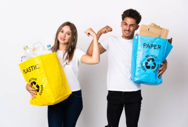 Beyaz arka planda izole edilmiş plastik ve kağıt dolu bir çanta tutan genç çift güçlü bir jest yapıyor.