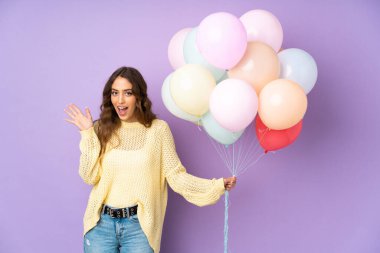 Mor arka planda bir sürü balon yakalayan genç bir kadın. Sürpriz bir yüz ifadesiyle.