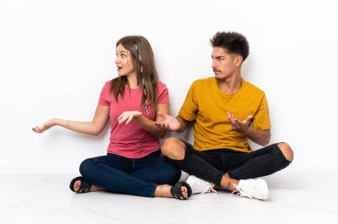 Yerde oturan genç çift, beyaz arka planda izole edilmiş, sürpriz bir ifadeyle yan tarafa bakıyor.