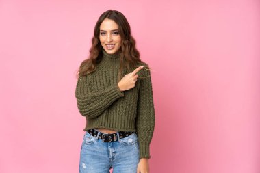 Soyutlanmış pembe arka planı olan genç bir kadın bir ürünü sunmak için kenarı işaret ediyor.