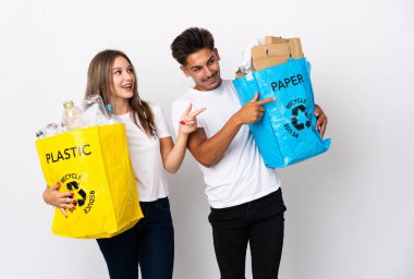 Beyaz arka planda izole edilmiş plastik ve kâğıt dolu bir çanta taşıyan genç çift, bir yandan işaret ediyor ve bir ürün sunuyor.