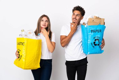 Beyaz arka planda izole edilmiş plastik ve kağıt dolu bir çanta taşıyan genç çift gülümserken yukarı bakıyor.