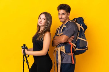 Sarı arka planda izole edilmiş büyük sırt çantalı genç dağcı çift kollarını yan pozisyonda tutarken gülümsüyor.