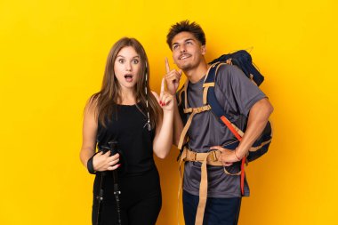Sarı arka planda izole edilmiş büyük sırt çantalı genç dağcı çift, bir fikir üzerinde hareket ediyorlar.