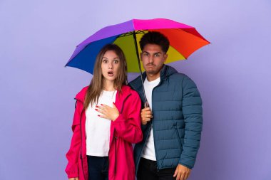 Mavi arka planda bir şemsiye tutan çift şaşırmış ve şok olmuş bir yüz ifadesine sahip.