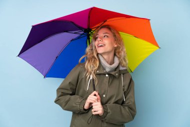 Mavi arka planda şemsiye tutan genç sarışın kadın mutlu bir ifadeyle izole edilmiş.
