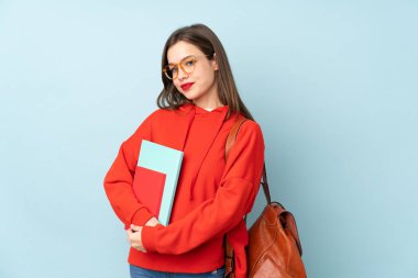 Mavi arka planda izole edilmiş kitapları tutan öğrenci kız yan tarafa bakıyor.