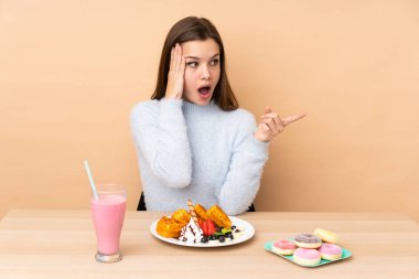 Bej arka planda izole edilmiş waffle yiyen genç kız şaşırdı ve parmağını yana çevirdi.
