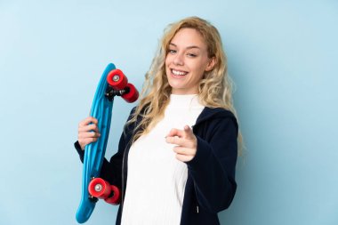 Genç sarışın kadın, mavi arka planda izole edilmiş, paten kayıyor ve ön tarafı işaret ediyor.