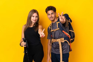 Sarı arka planda izole edilmiş büyük sırt çantalı genç dağcı çift gülümsüyor ve zafer işareti gösteriyor.