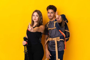 Sarı arka planda izole edilmiş büyük sırt çantalı genç dağcı çift parmağını kaldırıyor.