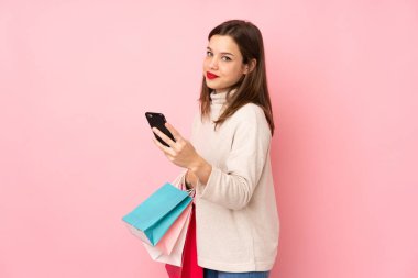 Pembe arka planda izole edilmiş genç bir kız elinde alışveriş çantaları ve bir arkadaşına cep telefonuyla bir mesaj yazıyor.