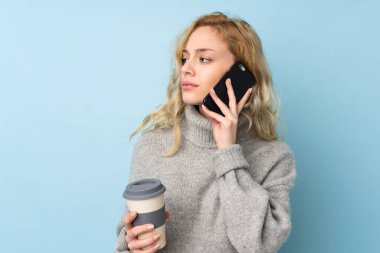 Mavi arka planda izole edilmiş süveter giyen genç sarışın bir kadın. Elinde kahve ve cep telefonu var.