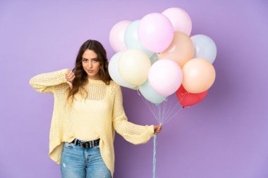 Mor arka planda bir sürü balon yakalayan genç bir kadın baş parmağını aşağı gösteriyor.