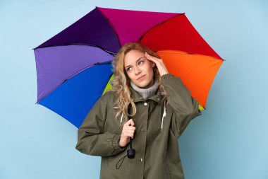 Mavi arka planda şemsiyesi olan genç sarışın kadının şüpheleri var ve kafası karışmış bir yüz ifadesi var.