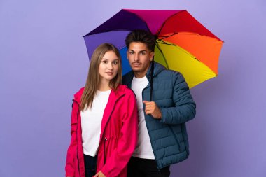 Mavi arka planda bir şemsiye tutan çift kollarını kalçalarına dayayıp gülümsüyor.