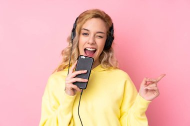 Pembe arka planda izole edilmiş sweatshirt giyen genç sarışın bir kadın cep telefonuyla müzik dinliyor ve şarkı söylüyor.