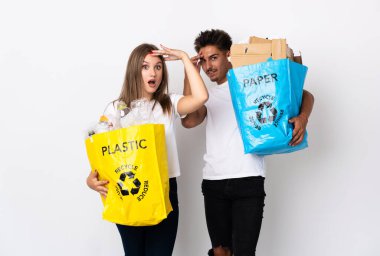 Beyaz arka planda plastik ve kağıt dolu bir çanta tutan genç çift yan tarafa bakarken sürpriz jest yapıyor.