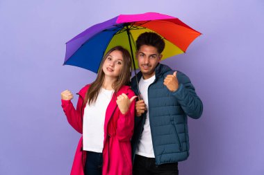 Mavi arka planda şemsiyesi olan çift iki eliyle baş parmak hareketi yapıyor ve gülümsüyor.