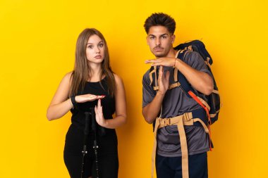 Sarı arka planda izole edilmiş büyük sırt çantalı genç dağcı çift bir gösteriyi durdurmak için el hareketi yapıyor.