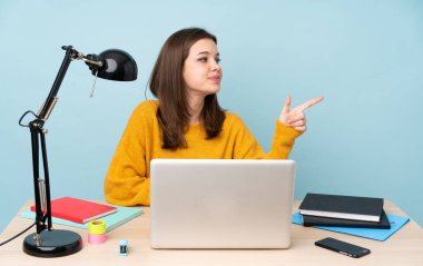 Öğrenci kız, mavi arka planda izole bir şekilde evinde çalışıyor. Parmağıyla bir ürünü gösteriyor.