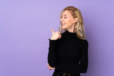 Mor arka planda izole edilmiş genç sarışın kadın bir ürünü sunmak için kenarı işaret ediyor.