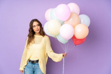 Mor arka planda bir sürü balon yakalayan genç bir kadın duruyor ve yan tarafa bakıyor.