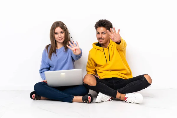 一对年轻的夫妇 带着笔记本电脑 坐在地板上 与白色背景隔离 幸福地数着四个手指 — 图库照片