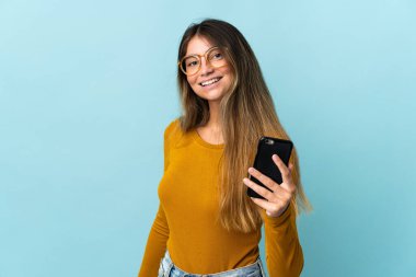 Mavi arka planda izole edilmiş, mutlu bir ifadeyle cep telefonu kullanan beyaz bir kadın.