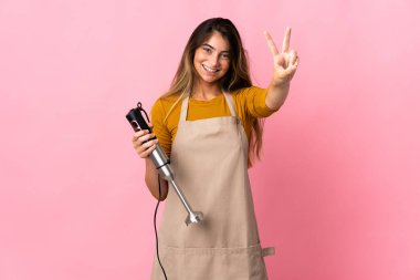 Genç aşçı kadın pembe arka planda izole edilmiş el blenderi kullanıyor. Gülümsüyor ve zafer işareti gösteriyor.