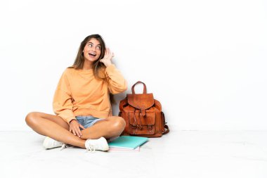 Yerde dizüstü bilgisayarlı genç bir bayan oturuyor. Beyaz arka planda izole edilmiş. Kulağına elini sürerek bir şeyler dinliyor.