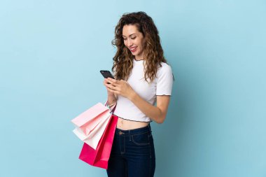 Kafkasyalı genç bir kadın mavi arka planda izole edilmiş. Elinde alışveriş poşetleri var ve bir arkadaşına cep telefonuyla bir mesaj yazıyor.