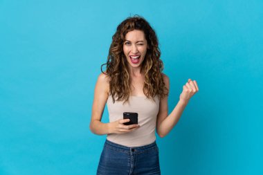 Genç beyaz kadın mavi arka planda izole edilmiş ve telefon zafer pozisyonunda.