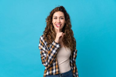 Mavi arka planda izole edilmiş beyaz bir kadın sessiz bir jest işareti gösteriyor. Parmağını ağzına sokuyor.