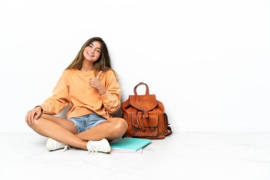 Yerde dizüstü bilgisayarlı genç bir bayan oturuyor ve beyaz arka planda izole edilmiş bir şekilde baş parmak işareti yapıyor.