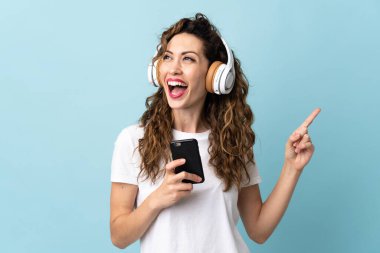 Genç beyaz kadın mavi arka planda izole edilmiş. Cep telefonuyla müzik dinliyor ve şarkı söylüyor.