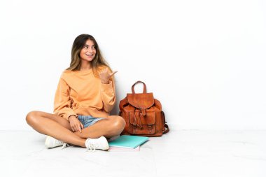 Genç bir bayan öğrenci dizüstü bilgisayarla yerde oturuyor beyaz arka planda izole edilmiş bir şekilde bir ürünü sunmak için kenarı gösteriyor.