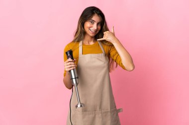 Pembe arka planda izole edilmiş el blenderi kullanan genç aşçı kadın telefon jesti yapıyor. Bana geri dönüş işareti ver.