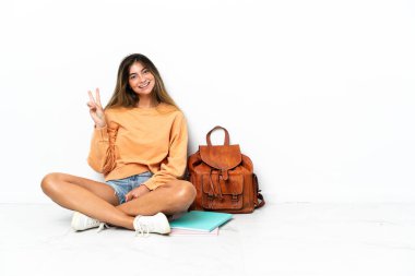 Yerde dizüstü bilgisayarlı genç bir bayan oturuyor. Beyaz arka planda izole edilmiş. Gülümsüyor ve zafer işareti gösteriyor.