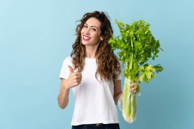 Mavi arka planda izole edilmiş kereviz tutan genç bir kadın. Başparmakları havada çünkü iyi bir şey oldu.